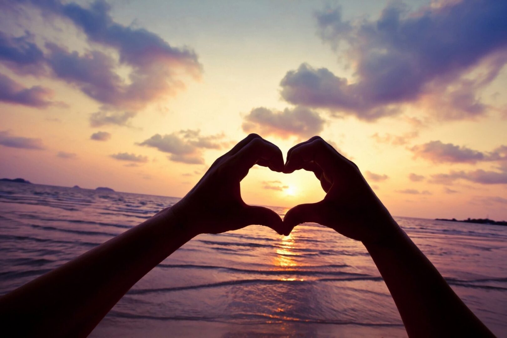 A person making a heart with their hands at sunset.